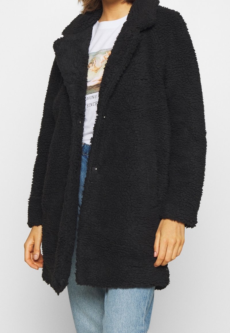 Abrigo negro y peludo confeccionado con una tela texturizada, que presenta un cuello con solapa en muesca, apertura frontal y mangas largas, combinado con una camiseta gráfica debajo.