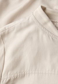 Beżowy materiał z gładkim wykończeniem, okrągłym dekoltem i płaskimi szwami. Tkanina wydaje się lekka i lekko błyszcząca, z miękką fakturą.