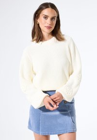 Frau mit schulterlangem braunem Haar, die einen weißen Strickpullover mit floralem Ellbogenpatch und einen blauen Cordrock trägt und nach vorne blickt.
