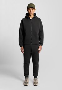 Svart zip-up hoodie med raglanärmar, matchad med svarta joggers. Bärs med en grön keps med en liten logga och beige sneakers.