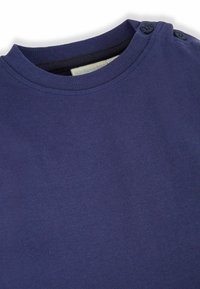 Námořnicky modrá košile s krátkým rukávem vyrobená z bavlny, s kulatým výstřihem a dvěma knoflíkovými detaily na levém rameni. Hladká textura.