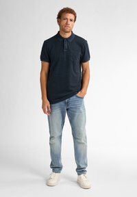 Polo cu dungi marine, cu mâneci scurte, cu buzunar pe piept, purtat cu blugi albaștri deschiși și adidași albi. Modelul stă în fața unui fundal deschis.