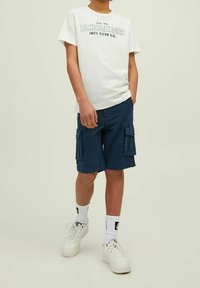 T-shirt bianca con testo nero, pantaloni cargo blu navy con due tasche laterali, sneaker bianche e calze bianche con dettagli neri.