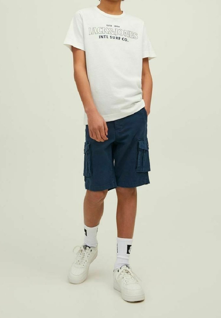 T-shirt bianca con testo nero, pantaloni cargo blu navy con due tasche laterali, sneaker bianche e calze bianche con dettagli neri.