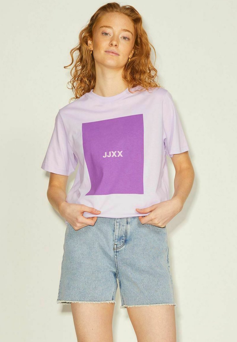 Camiseta de algodón lila de mangas cortas con un gran gráfico cuadrado púrpura y el texto blanco "JJXX", combinada con pantalones cortos de mezclilla azul claro.
