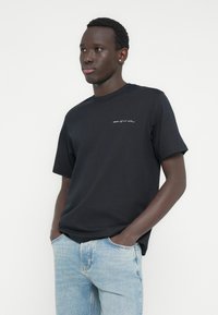 Μαύρο βαμβακερό T-shirt με κοντά μανίκια, στρογγυλή λαιμόκοψη και λευκό κεντητό κείμενο στο αριστερό στήθος. Φορεμένο με ανοιχτό μπλε τζιν παντελόνι.