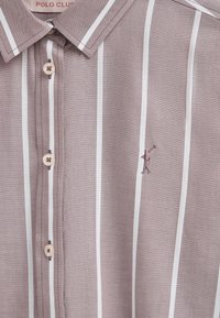 Korte mouw, lichtbruine blouse met een kraag en verticale witte strepen. Voorzien van een knoopsluiting en een klein geborduurd figuurtje op de borst.