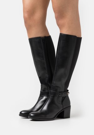 Botas de cuero negro brillante hasta la rodilla con cremallera lateral, detalle de correa alrededor del tobillo y tacones gruesos, usadas sobre piernas desnudas contra un fondo blanco.