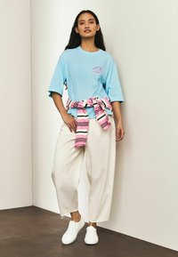 Svetlo modra oversized majica z roza grafiko, kombinirana z belimi širokimi hlačami in roza črtastim puloverjem, zvezanim okoli pasu. Bele superge.