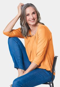Oranges ¾-Ärmeloberteil mit drapiertem Ausschnitt, kombiniert mit blauen Jeans. Das Motiv sitzt und zeigt die lockere Passform und die glatte Textur des Oberteils.
