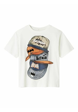 T-shirt blanc avec un motif représentant des casquettes de baseball empilées en beige, orange et bleu, avec le texte « BATMAN » et des symboles de chauve-souris.