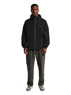 Mann steht und trägt eine schwarze Kapuzenjacke mit Reißverschluss, graue Hose sowie graue und weiße Turnschuhe vor weißem Hintergrund.