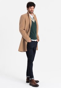 Beige ullkappa, grön v-ringad tröja, vit skjorta med krage, mörka jeans, bruna läderankelstövlar; modell står med händerna i fickorna.