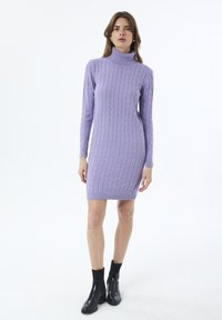 Frau steht in einem lilafarbenen, strukturierten Turtleneck-Pulloverkleid mit schwarzen Knöchelstiefeln vor einem weißen Hintergrund.