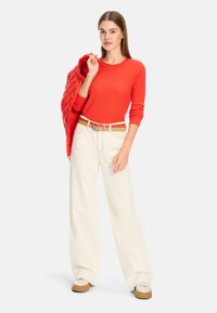 Roter gerippter Pullover, cremefarbene weite Hose, beiger Gürtel mit einer Schnalle. Model hält eine rote gesteppte Jacke. Texturiertes Material, lockere Passform.