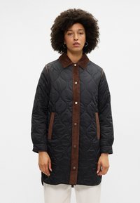 Kvadreast plašč v črni barvi z rjavim žametnim ovratnikom in gumbi, ki ima pokončne rjave poudarke in stranske žepke. Gladka tekstura po vsej površini.