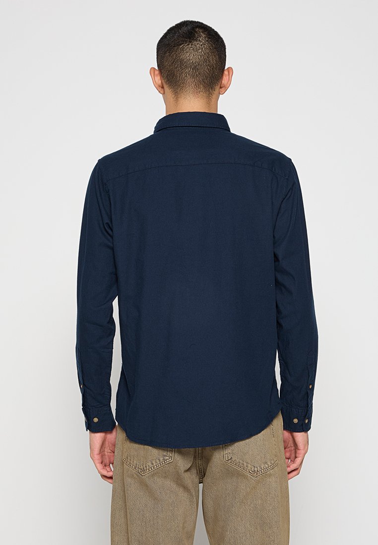 Camisa de botones de manga larga azul marino con corte recto, con una sola yugo trasero y dobladillo redondeado, que presenta puños con botones y una textura suave.