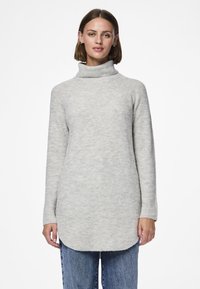 Sieviete, kas valkā garu, gaiši pelēku augstu apkaklīti ar džinsiem, stāv pret vienveidīgu baltu fonu.