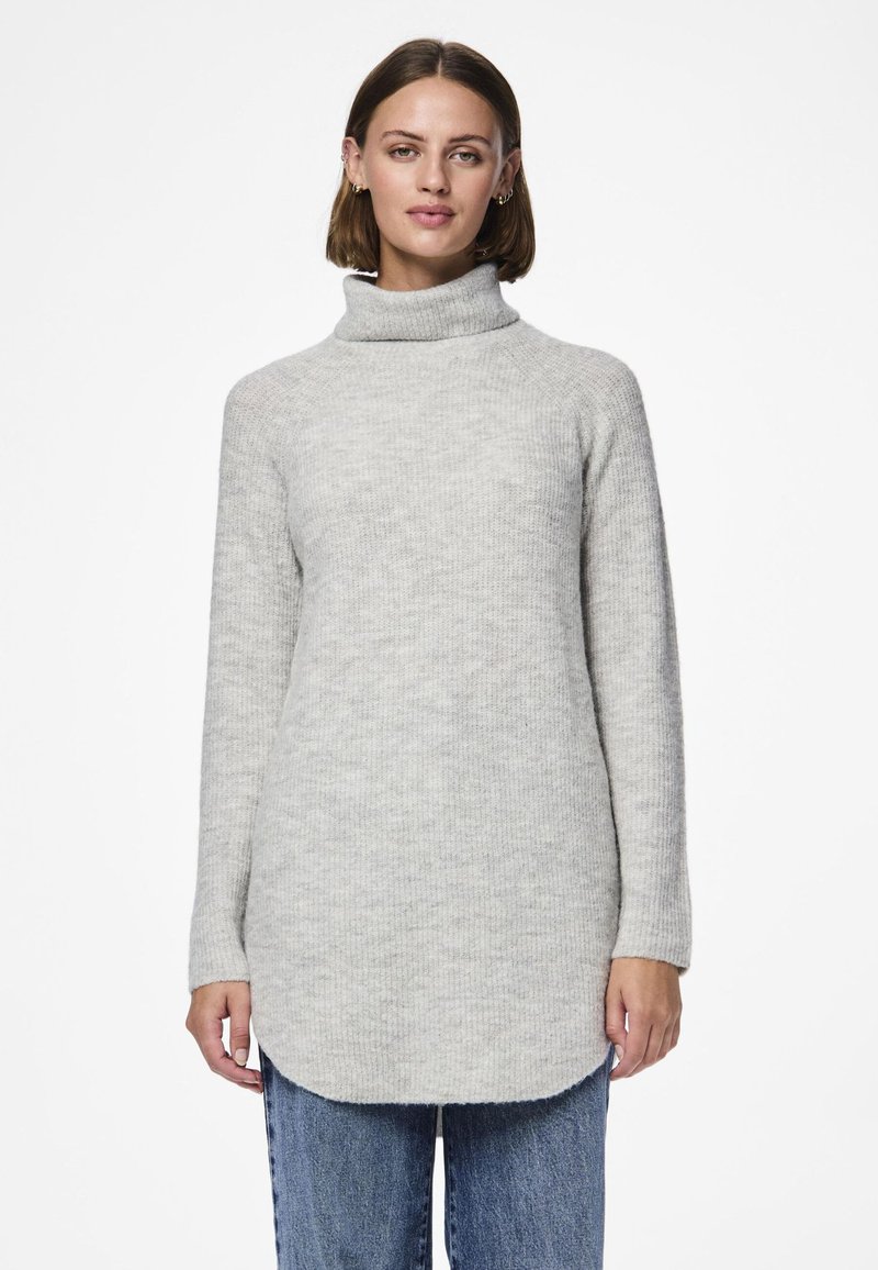 Sieviete, kas valkā garu, gaiši pelēku augstu apkaklīti ar džinsiem, stāv pret vienveidīgu baltu fonu.