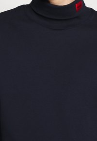 Námořnický rolák vyrobený z jemné tkaniny, s vysokým límcem a červenou štítkovou nášivkou na krku. Hladká textura a přiléhavý střih.