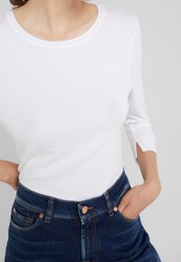 Kvinna iklädd en vit långärmad skjorta med liten slits i ärmarna och mörkblå högmidjade jeans, med ena handen i fickan.