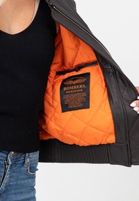 Czarna skórzana kurtka bomberka z pikowanym pomarańczowym podszyciem. Posiada ściągacze na mankietach oraz metkę z napisem "BOMBERS HERITAGE" wszytą w podszycie.