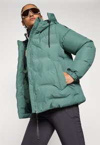 Person som bär en överdimensionerad grön pufferjacka, mörka leggings och stora beige solglasögon, poserar med en hand i jackfickan mot en vit bakgrund.