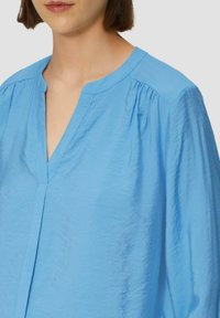 Lichtblauwe blouse met een geknipte kraag, geplooide details op de schouders en een licht gekreukt textuur. Heeft een knoopsluiting aan de voorkant.