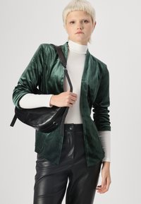 Zelený sametový blejzer s dlhými rukávmi, skombinovaný s bielym rolákom. Čierne kožené nohavice a čierna crossbody taška. Hladká štruktúra.