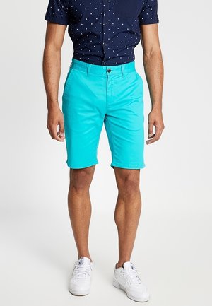 Homme portant un short turquoise vif arrivant aux genoux, une chemise à manches courtes bleu marine avec un motif blanc, et des baskets blanches.