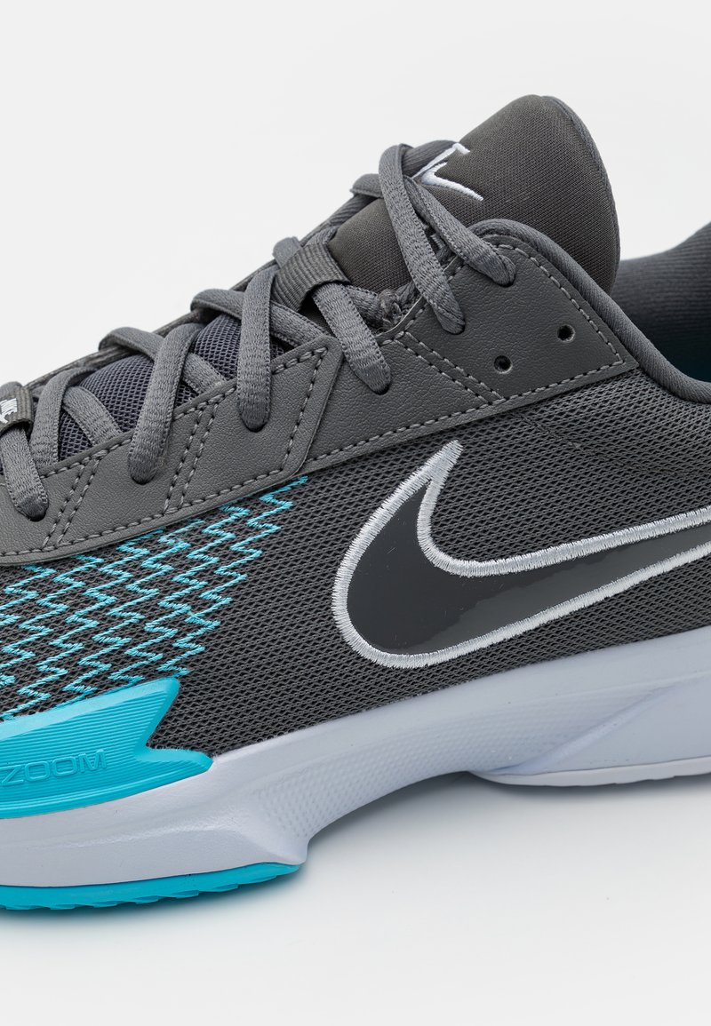 Zapatilla Nike con parte superior de malla gris, acentos turquesa y un swoosh negro brillante. Presenta una lengüeta acolchada y cordones texturizados.