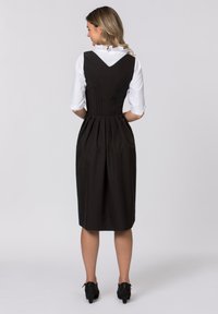 Schwarzes knielanges Kleid mit einem taillierten Oberteil, V-Ausschnitt hinten und plissiertem Rock. Trägt eine weiße Bluse mit einer Rüsche am Kragen. Schwarze Schuhe.