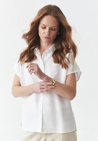 Camicia bianca a maniche corte con bottoni realizzata in tessuto leggero, con una vestibilità morbida. Presenta un colletto e spacchi laterali per una maggiore libertà di movimento.