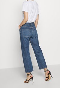 Person klädd i blå jeans med hög midja, vit t-shirt och svarta högklackade sandaler med remmar, stående mot en enkel vit bakgrund.