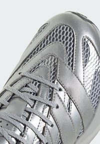 Stříbrná sportovní bota s síťovaným svrškem a metalickými akcenty, texturovanými panely a plochými tkaničkami. Zaoblená špička a polstrovaná podešev.