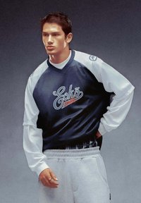 Maglia a maniche lunghe in rete navy con maniche bianche, con logo "Eckō". Abbinata a pantaloni sportivi grigi. Tessuto testurizzato e vestibilità atletica.