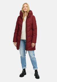 Manteau rembourré bordeaux matelassé avec capuche ; doté d'une fermeture éclair et de poches latérales. Porté sur un pull léger et un jean bleu clair.