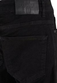 Melns džinsu audums ar ādas jostas tagu, uz kura rakstīts "PREMIUM 2Y", redzamām diega šuvēm un aizmugurējās kabatas dizainu.