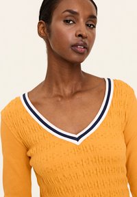 Gelber Strickpulli mit tiefem V-Ausschnitt und geripptem Textur, mit einem kontrastierenden Kragen in Weiß und Marineblau mit Streifen.