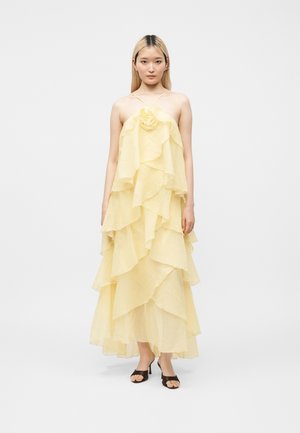 Jeune femme portant une robe à couches, jaune pâle, longueur cheville, à fines bretelles, avec des talons noirs à bout ouvert, debout devant un fond uni.