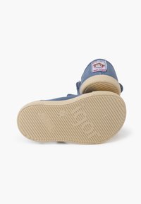 Zapatilla de lona azul con suela de goma marrón. Presenta una etiqueta colorida en la parte superior y una suela texturizada con patrones de agarre. Fabricada en España.