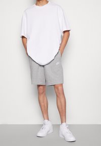 Bílé oversized tričko, šedé atletické kraťasy s logem Nike a bílé tenisky s vysokým střihem. Materiál působí měkce a volně.