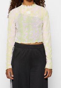 Frau trägt ein kurzes, langärmliges Oberteil mit einem hellgrünen und pinken Schlangenmuster und Adidas-Logo, kombiniert mit schwarzen Adidas-Hosen mit weißen Streifen.