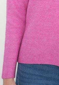 Suéter de malha em rosa brilhante, com mangas longas e textura de canelado. Combinado com um tecido de denim, destacando o contraste vibrante de cores.