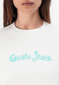 Balta sportinė palaidinė su turkio spalvos "Guess Jeans" logotipu, kuris turi žaismingą šriftą ir gėlių detales, pagaminta iš minkštos, tekstūruotos medžiagos.