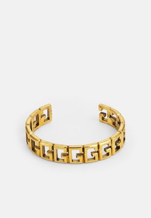 Bransoletka w kolorze złota z otwartym mankietem, zgeometryzowanymi prostokątnymi ogniwami tworzącymi ciągły wzór na białym tle.