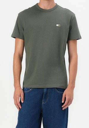 Mężczyzna w prostym zielonym T-shircie z krótkim rękawem z małym białym i czarnym logo na lewej piersi oraz w niebieskich dżinsach.