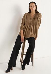 Brun plisseret blouse med V-hals og elastiske ærmer, parret med sorte vidde bukser og sorte ankelstøvler med hæl, siddende på en skammel.