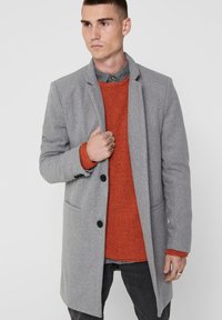 Grauem Wollmantel mit schwarzen Knöpfen, begleitet von einem orangefarbenen strukturierten Pullover und einem blauen Hemd darunter.