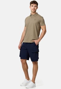 Oliwkowa koszulka polo z kołnierzykiem, zestawiona z granatowymi szortami bojówkami z wieloma kieszeniami. Model nosi białe tenisówki.
